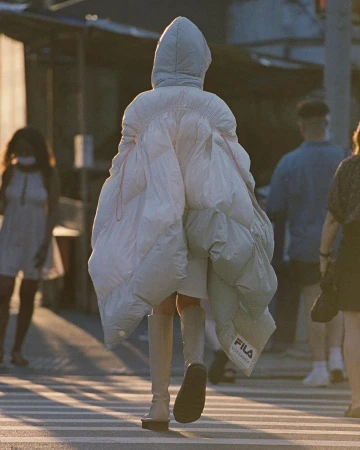 Person wearing oversized white puffer coat crossing a street in an urban setting with other people visible in the background.