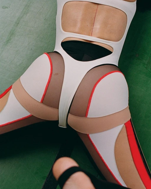 Close-up studio shot of a person lying on a green surface, wearing a white cut-out bodysuit with beige and red trim, and black strappy heels.