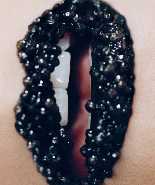 Close-up of an adult female's mouth with black beads applied to lips and visible white teeth. Studio lighting and skin detail are present.
