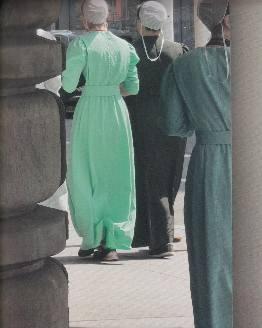 Three women in traditional dresses and head coverings stand on a sidewalk near a carved stone pillar, capturing a blend of fashion and culture.