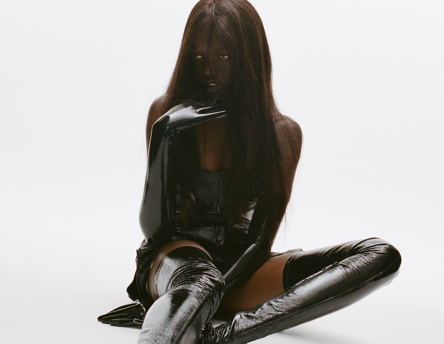Portrait of a Black woman with long straight hair, wearing a black corset top and shiny black thigh-high boots, posing in a studio.