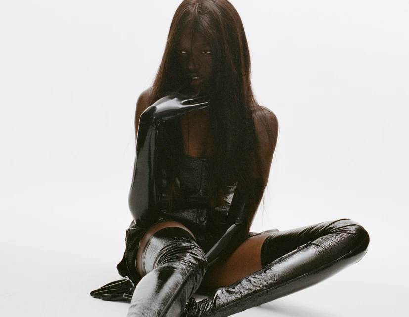 Portrait of a Black woman with long straight hair, wearing a black corset top and shiny black thigh-high boots, posing in a studio.