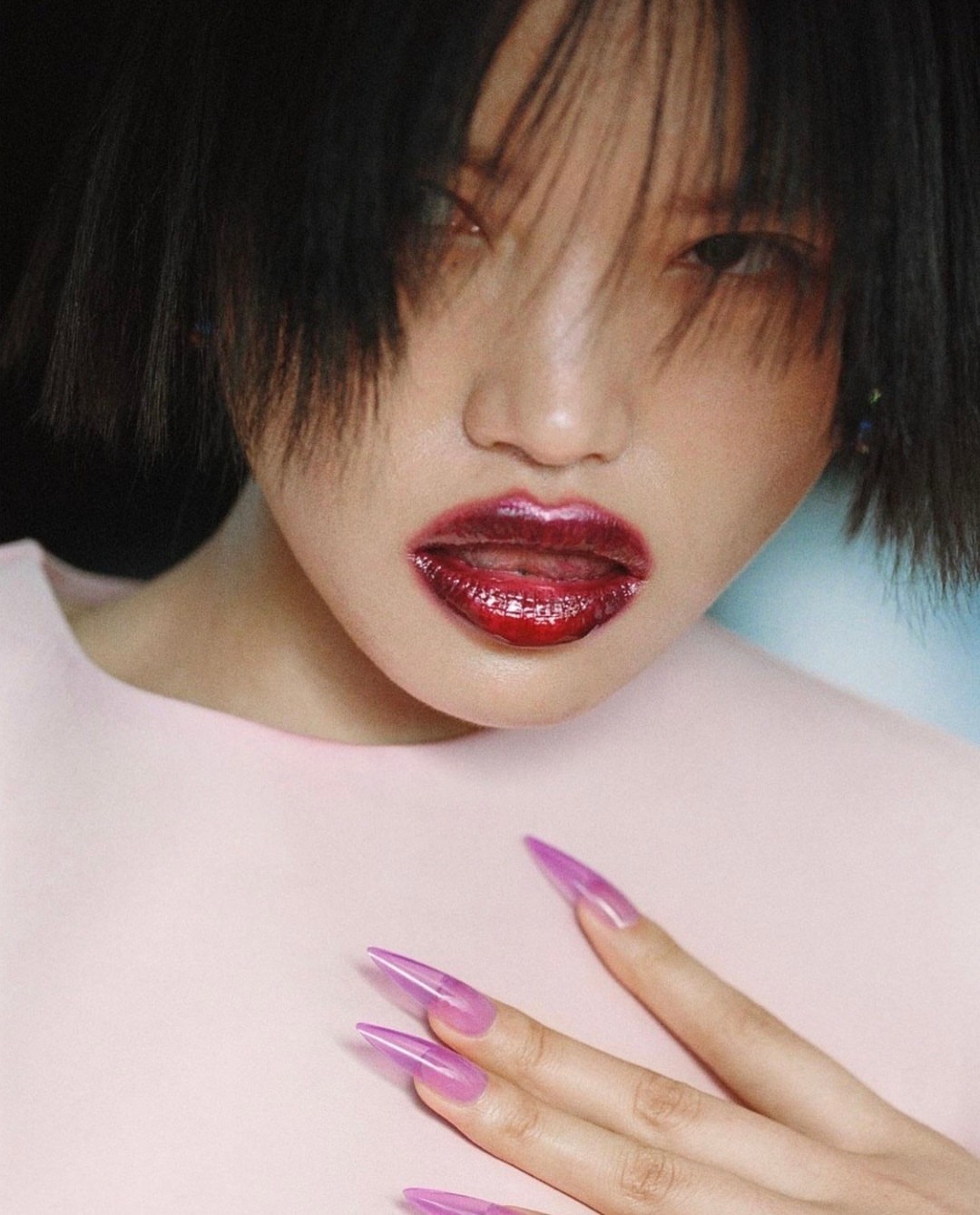 Close-up portrait of a woman with pink-tinted, glossy lips and clear, pointy acrylic nails against a soft pink backdrop.
