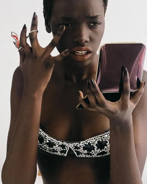 Close-up shot of a Black woman in jeweled bra, holding a compact mirror, touching her face, and showcasing long, black, almond-shaped nails.
