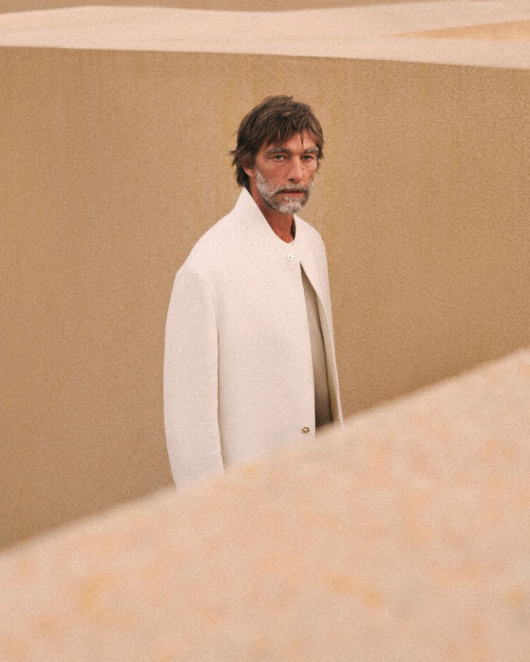 Mature adult man stands in front of tan walls wearing a light coat. Focus is on the male figure in a minimalist setting.