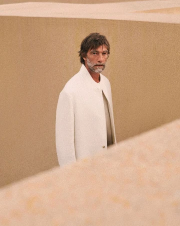 Mature adult man stands in front of tan walls wearing a light coat. Focus is on the male figure in a minimalist setting.