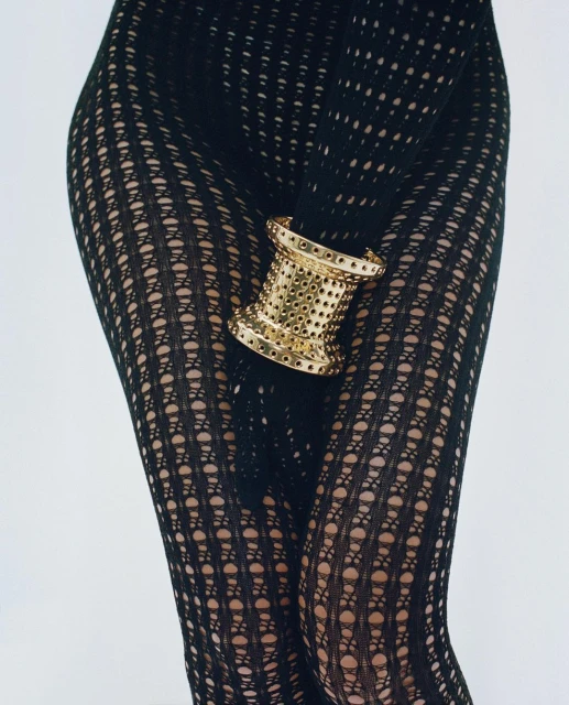 Close-up shot of a person wearing black patterned tights with a gold, drum-shaped cuff bracelet around their thigh on white.