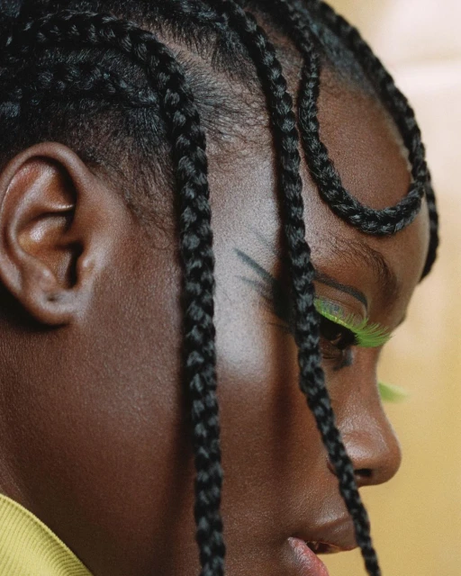 Close-up of a young Black adult female with braided hair and green eye makeup against a warm yellow background.