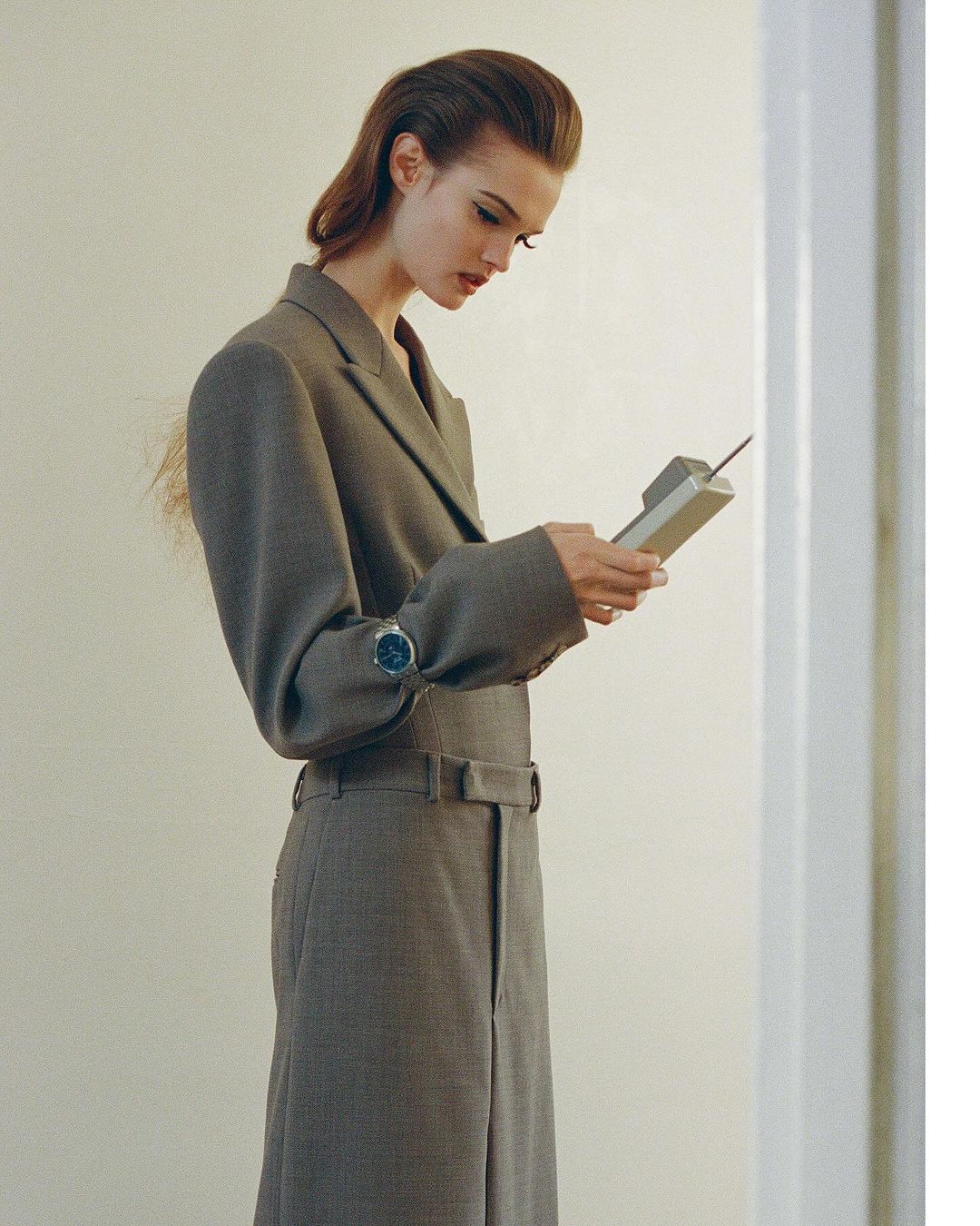 Elegant woman in a grey suit holds a vintage brick cell phone in an indoor setting, showcasing fashion and technology elements.