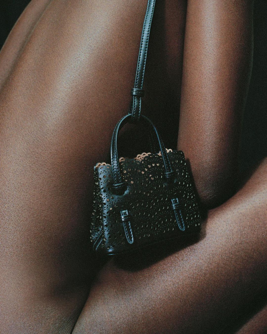Close-up of a black leather purse with hardware detail, hanging against a blurred brown skin background, studio shot.