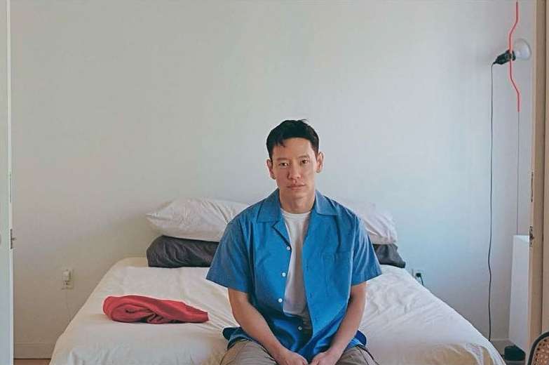 Adult male sits on a bed in a bright indoor space with white walls, wearing a blue button-down shirt and khaki pants. Natural light.