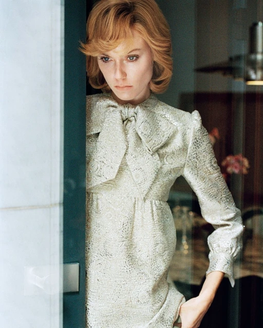 Blonde woman in patterned silver dress with bow poses next to doorway. Indoor shot with soft lighting and neutral tones.
