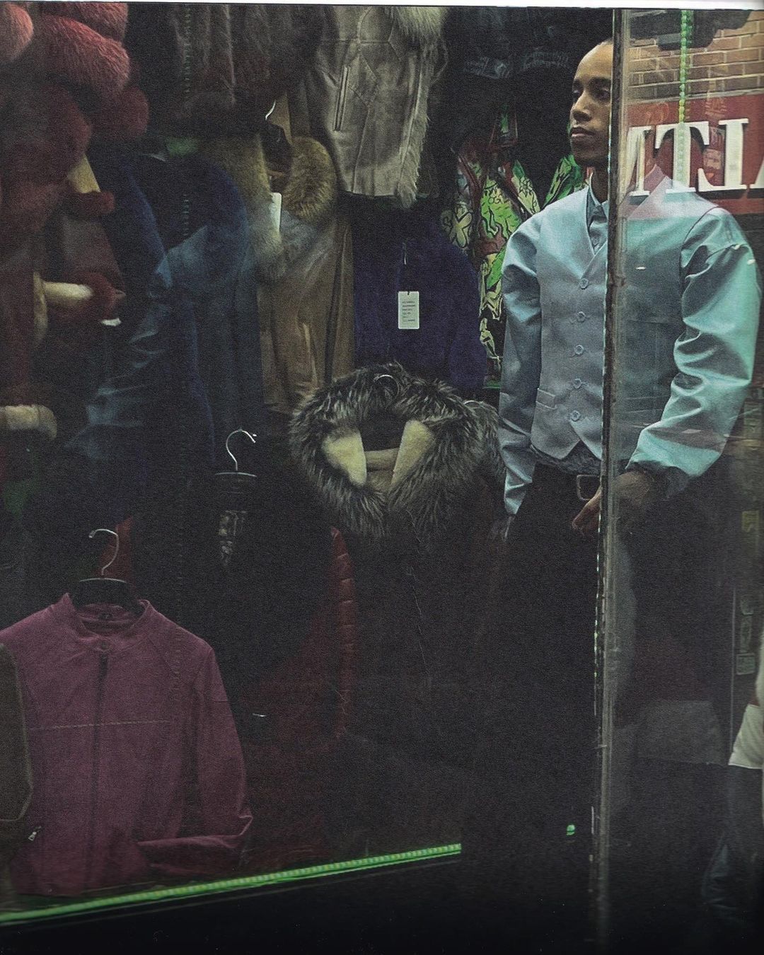 Bald adult male in a light blue vest and black pants standing in front of a clothing store window displaying fur coats and jackets.
