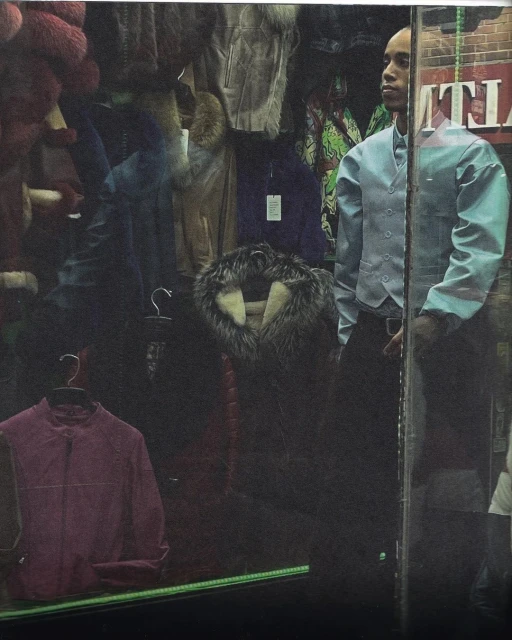 Bald adult male in a light blue vest and black pants standing in front of a clothing store window displaying fur coats and jackets.