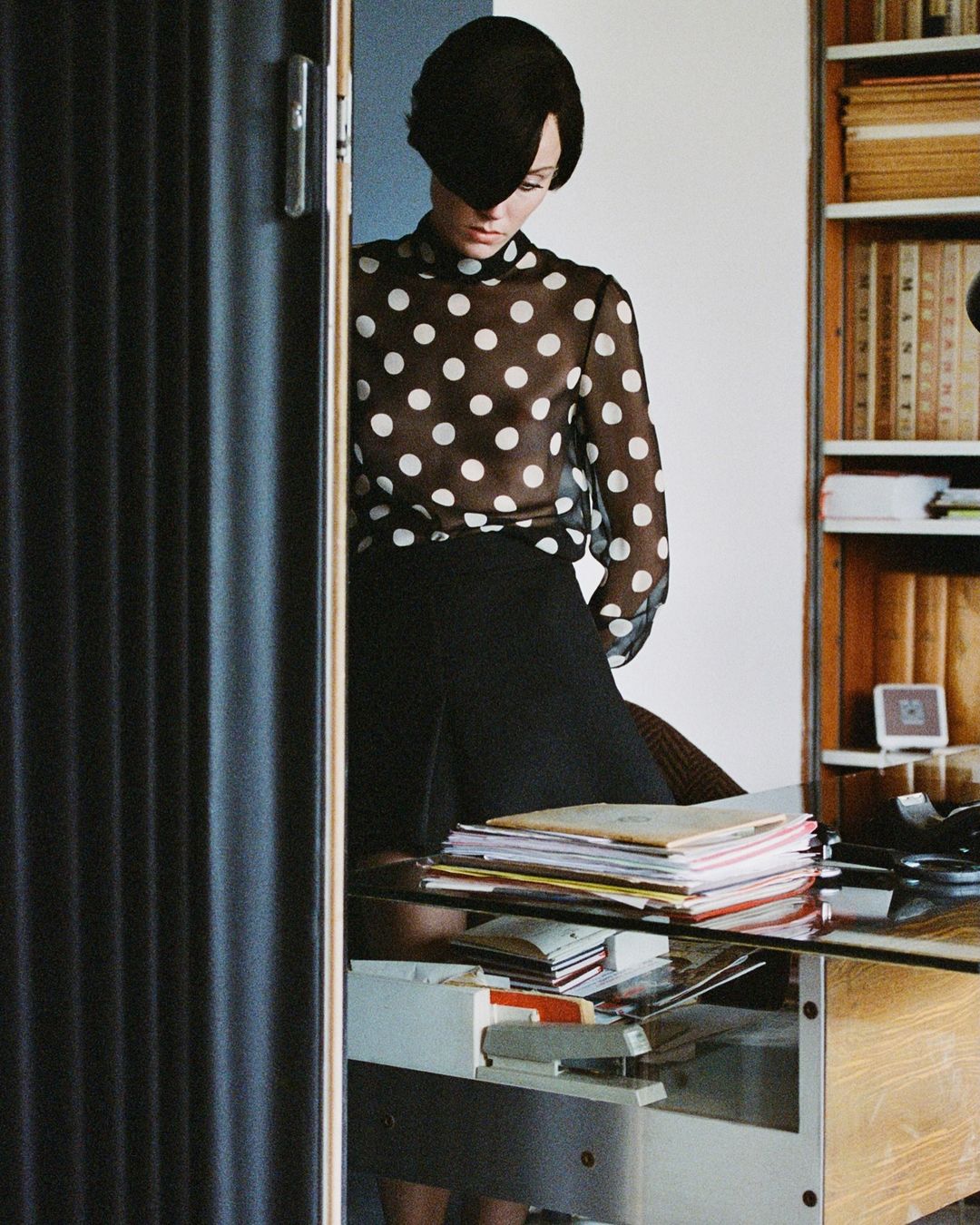Fashionable woman in polka dot blouse and black hat poses near a desk in a bright room with a bookshelf and interior doors.