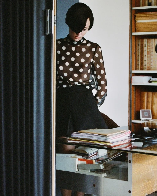 Fashionable woman in polka dot blouse and black hat poses near a desk in a bright room with a bookshelf and interior doors.