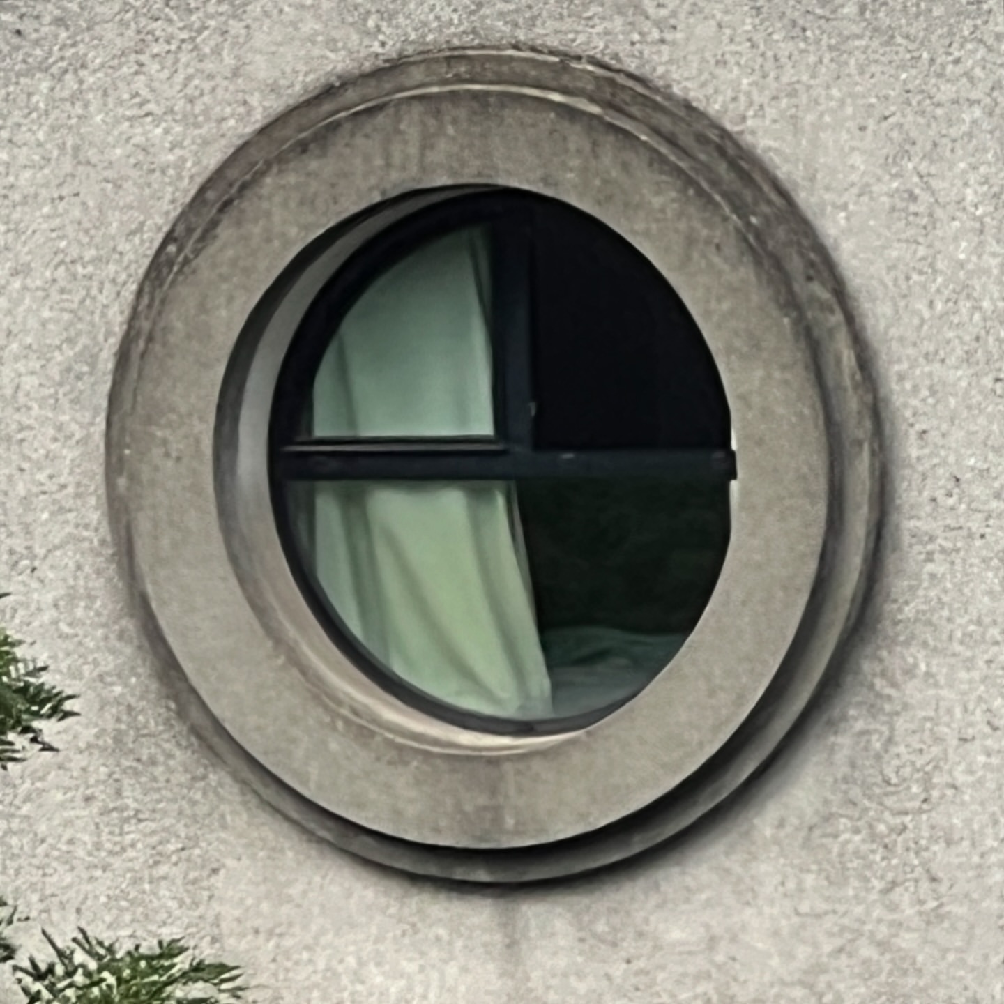 A round window in a grey stone wall offers a glimpse of a green curtain, casting a soft, diffused light within an interior space.