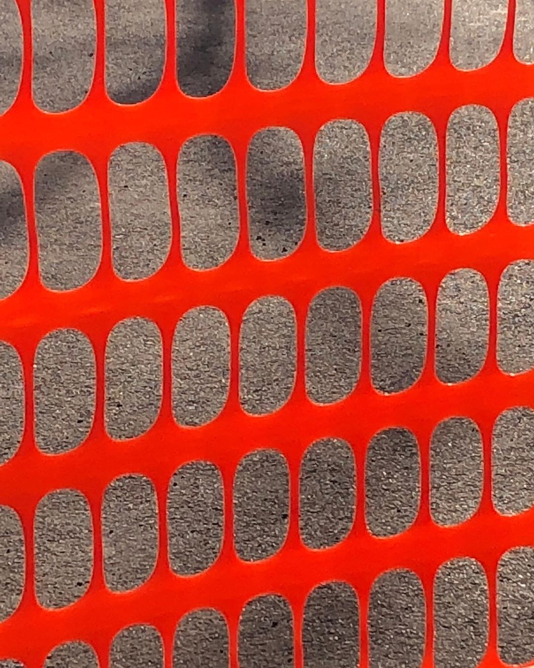 Safety fence close-up showing an orange plastic mesh with oval openings, casting shadows on the asphalt surface behind.