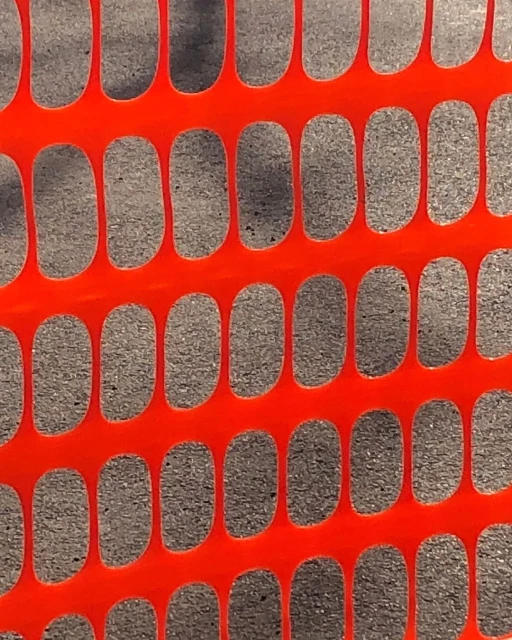Safety fence close-up showing an orange plastic mesh with oval openings, casting shadows on the asphalt surface behind.