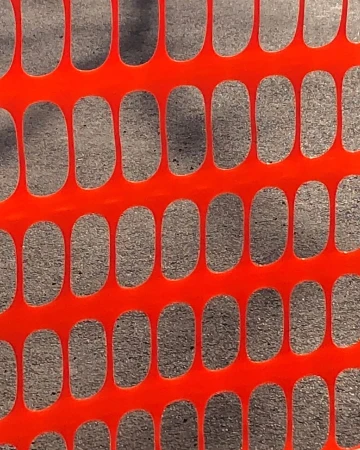 Safety fence close-up showing an orange plastic mesh with oval openings, casting shadows on the asphalt surface behind.