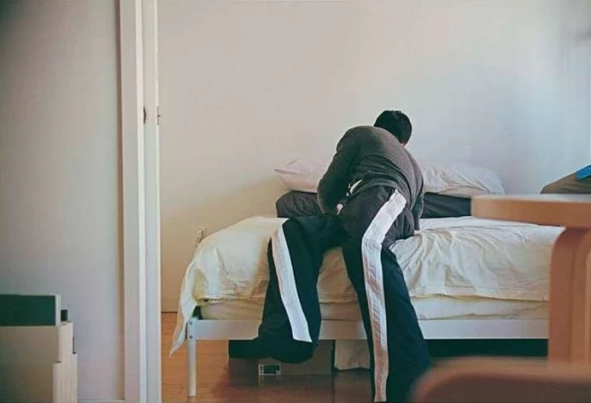 A person sits on a white bed in an indoor setting. The individual wears a grey shirt and dark pants with white stripes, facing away from the camera.