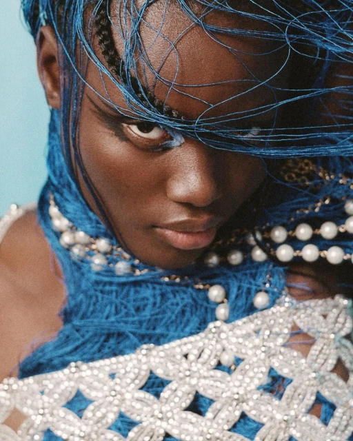 Close-up portrait of a Black female with blue beaded hair, pearl necklace, and silver embellished clothing against a blue background.