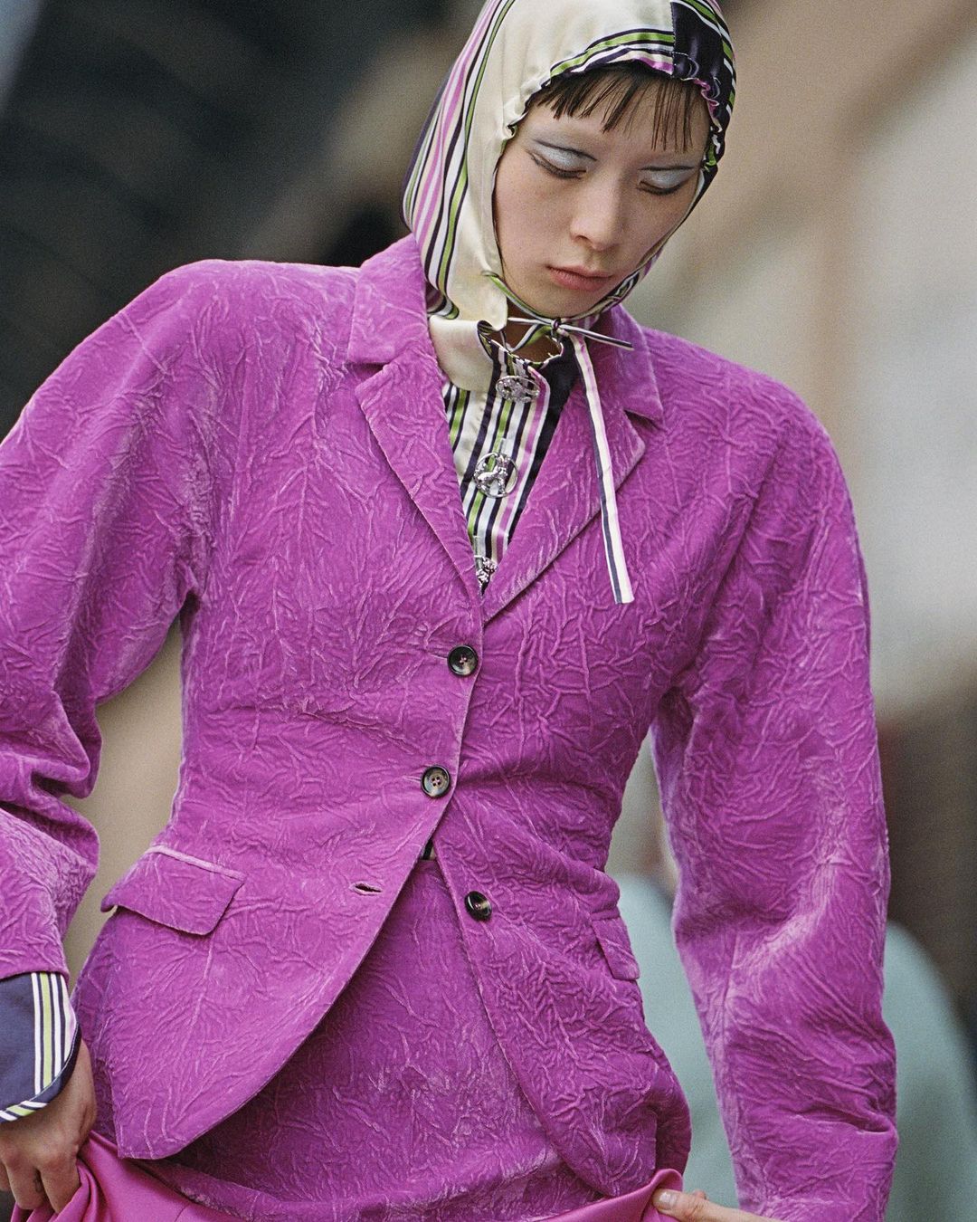 Runway fashion shot featuring a model in a purple blazer and skirt, with a striped headscarf, makeup, and jewelry, against an urban backdrop.