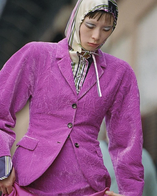 Runway fashion shot featuring a model in a purple blazer and skirt, with a striped headscarf, makeup, and jewelry, against an urban backdrop.