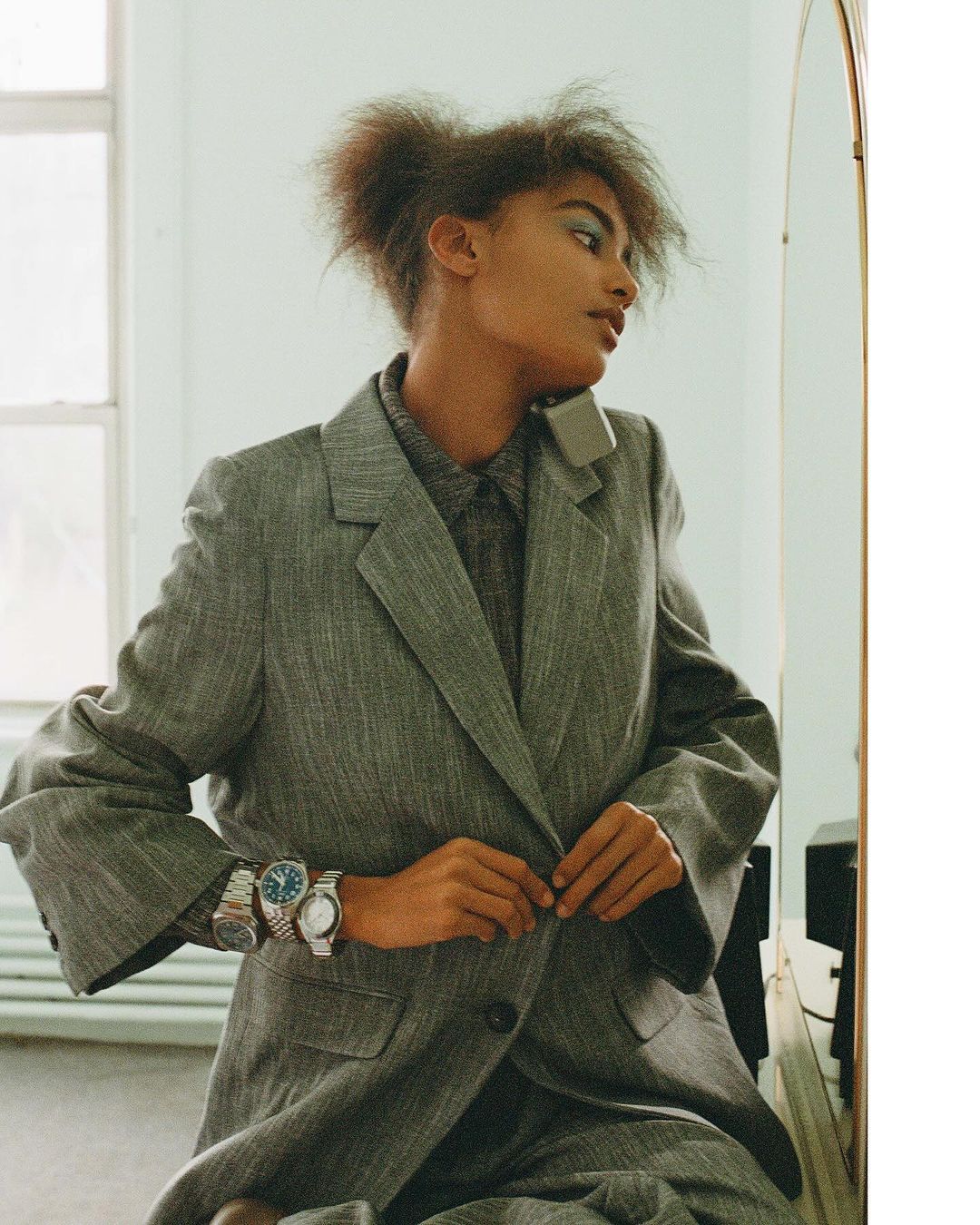 Woman in oversized grey suit fastens buttons, wearing multiple wristwatches, and reflecting in a nearby mirror in a bright indoor setting.