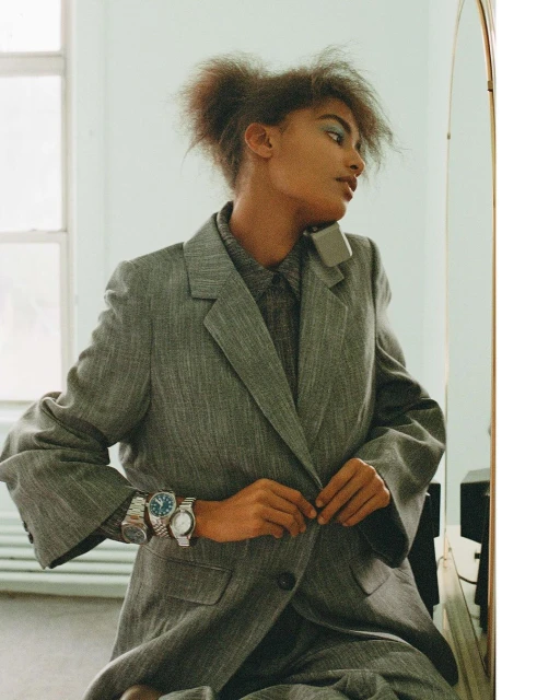 Woman in oversized grey suit fastens buttons, wearing multiple wristwatches, and reflecting in a nearby mirror in a bright indoor setting.