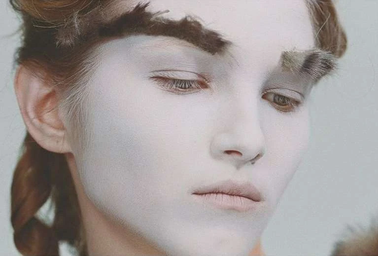 Close-up of a young woman with pale skin and dark hair, featuring dramatic makeup with heavy, feathered eyebrows and downward gaze.