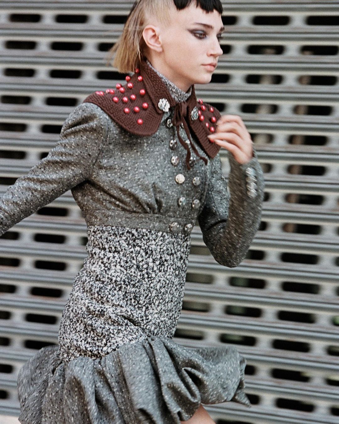A stylish young woman in a tweed jacket and dress poses in front of a corrugated metal wall, showcasing fashion-forward design.
