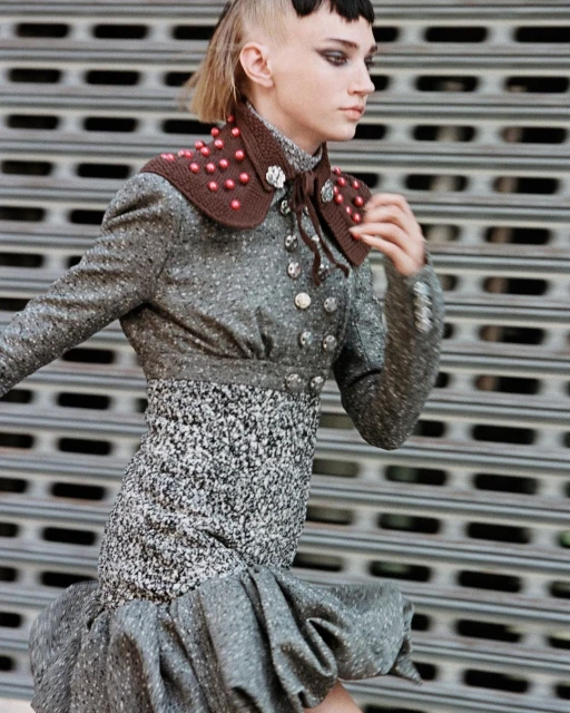 A stylish young woman in a tweed jacket and dress poses in front of a corrugated metal wall, showcasing fashion-forward design.
