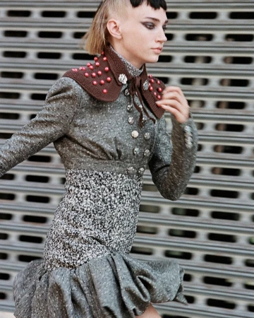 A stylish young woman in a tweed jacket and dress poses in front of a corrugated metal wall, showcasing fashion-forward design.