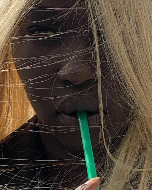 Close-up of an adult female drinking from a green straw, with blonde hair partially covering her face, set against a neutral background.