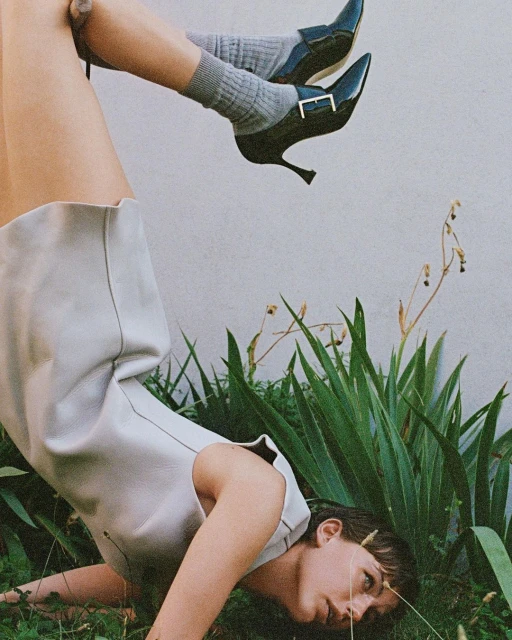 A woman in a light-colored dress and black high heels poses upside down outdoors among green plants on a sunny day, creating a unique fashion statement.