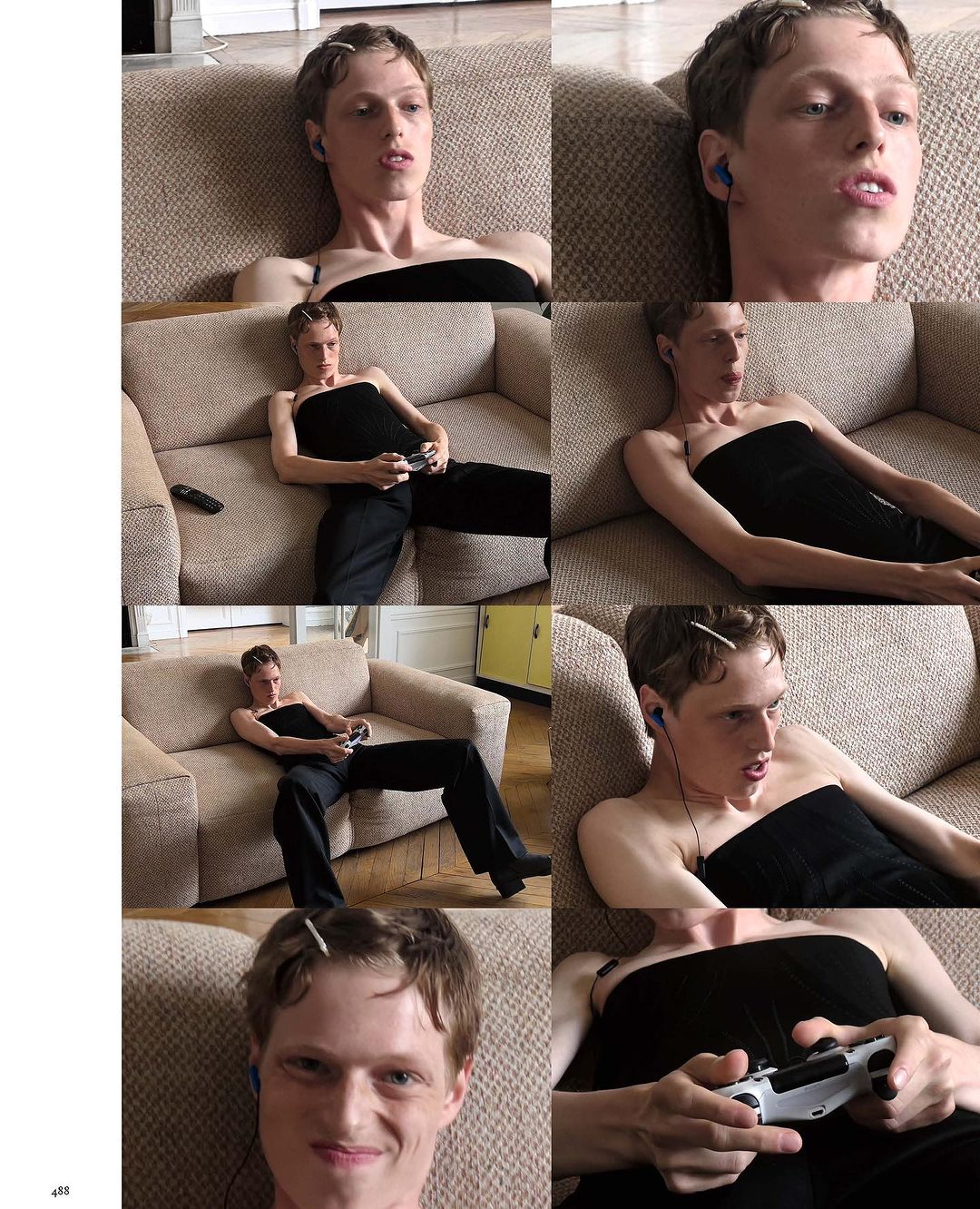 A person with short brown hair, wearing a black strapless top and dark pants, sits on a couch and plays a video game with a white controller.