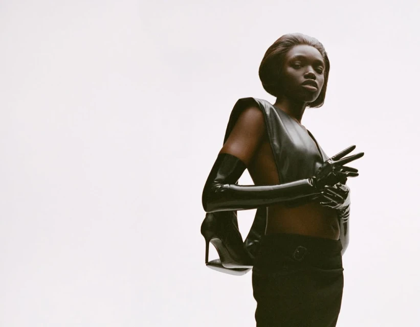 Elegant Black woman poses in a sleeveless black leather top, black skirt, long gloves, and heels against a bright white backdrop.