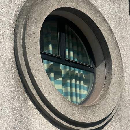 Exterior architectural shot of a circular window with blue and white striped curtains inside, set in a textured concrete wall.