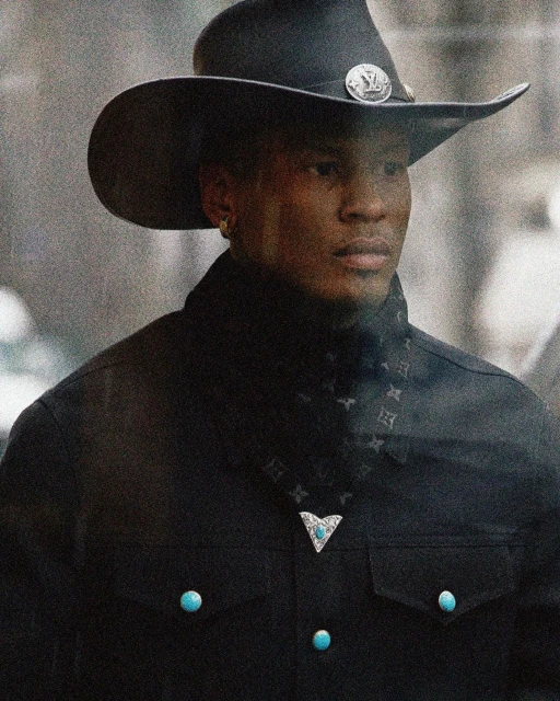 Man wearing a black cowboy hat, patterned bandana, and jacket with turquoise accents against an out-of-focus background.