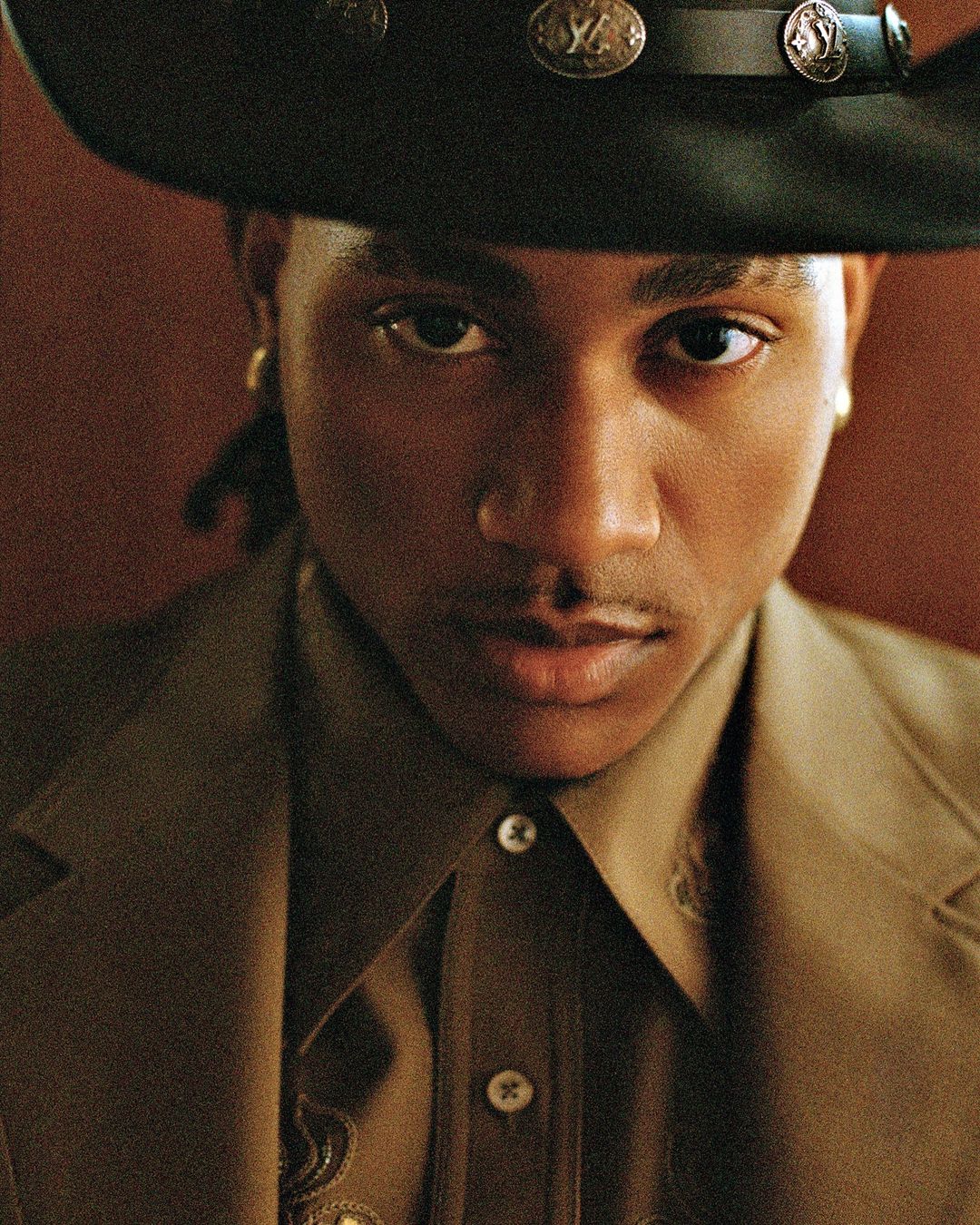 Close-up portrait of a young adult male with dark skin wearing a cowboy hat, button-down shirt, and blazer against a dark background.