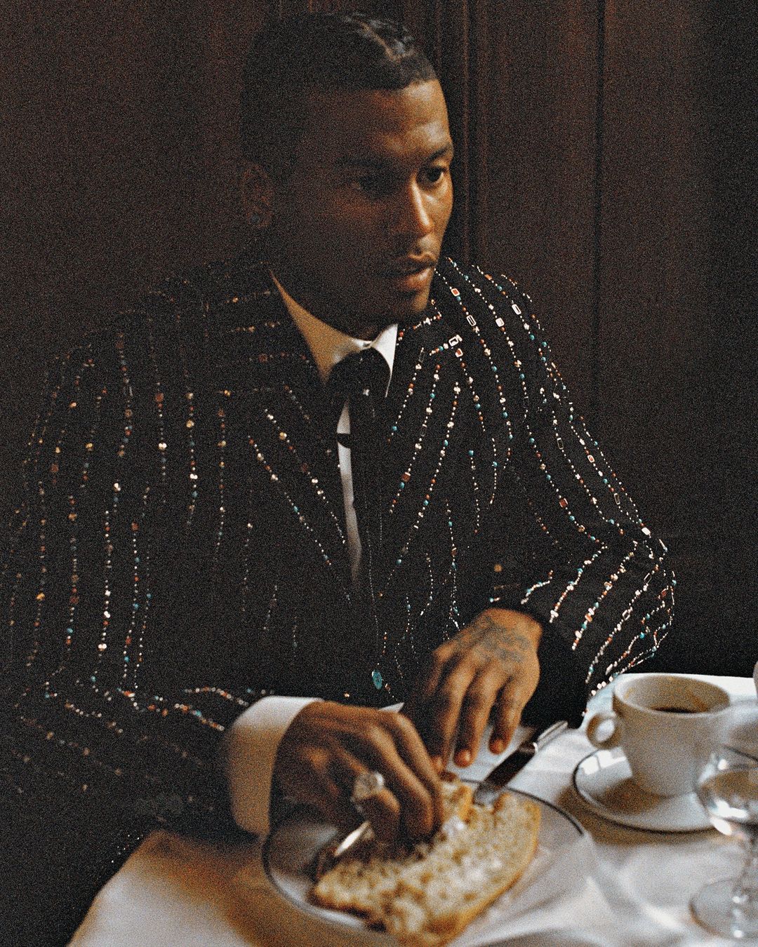 An African-American man in a sequined blazer sits at a table with a plate of bread, a cup of coffee, and a wine glass in a dimly lit room.