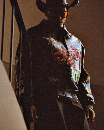 Man wearing a cowboy hat, tinted sunglasses, and an embellished dark leather jacket standing near a staircase.