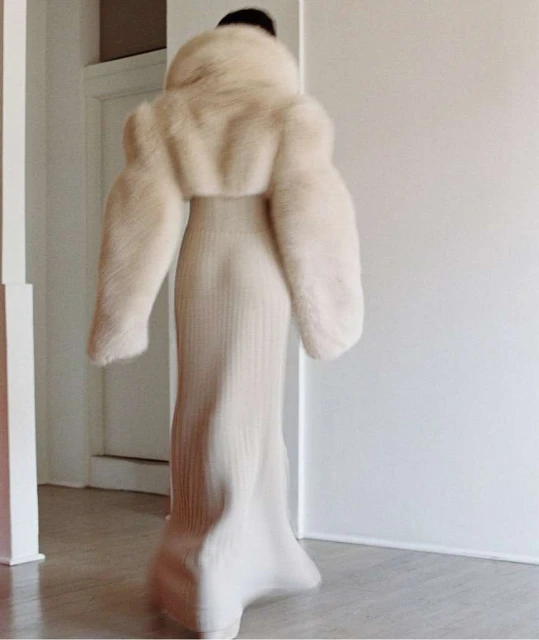 Woman wearing a long white ribbed dress and cream-colored fur jacket stands in a minimalist white interior with a doorway in the background.