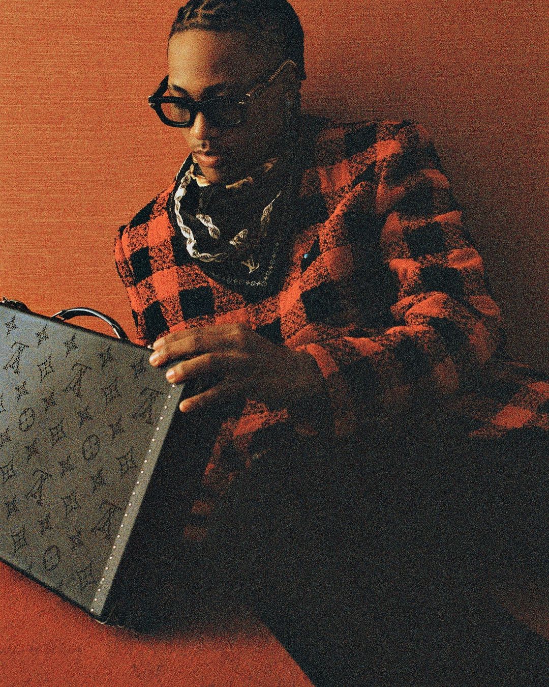 Young adult male with sunglasses poses indoors wearing a red plaid jacket, bandana, and holding a monogrammed Louis Vuitton case.
