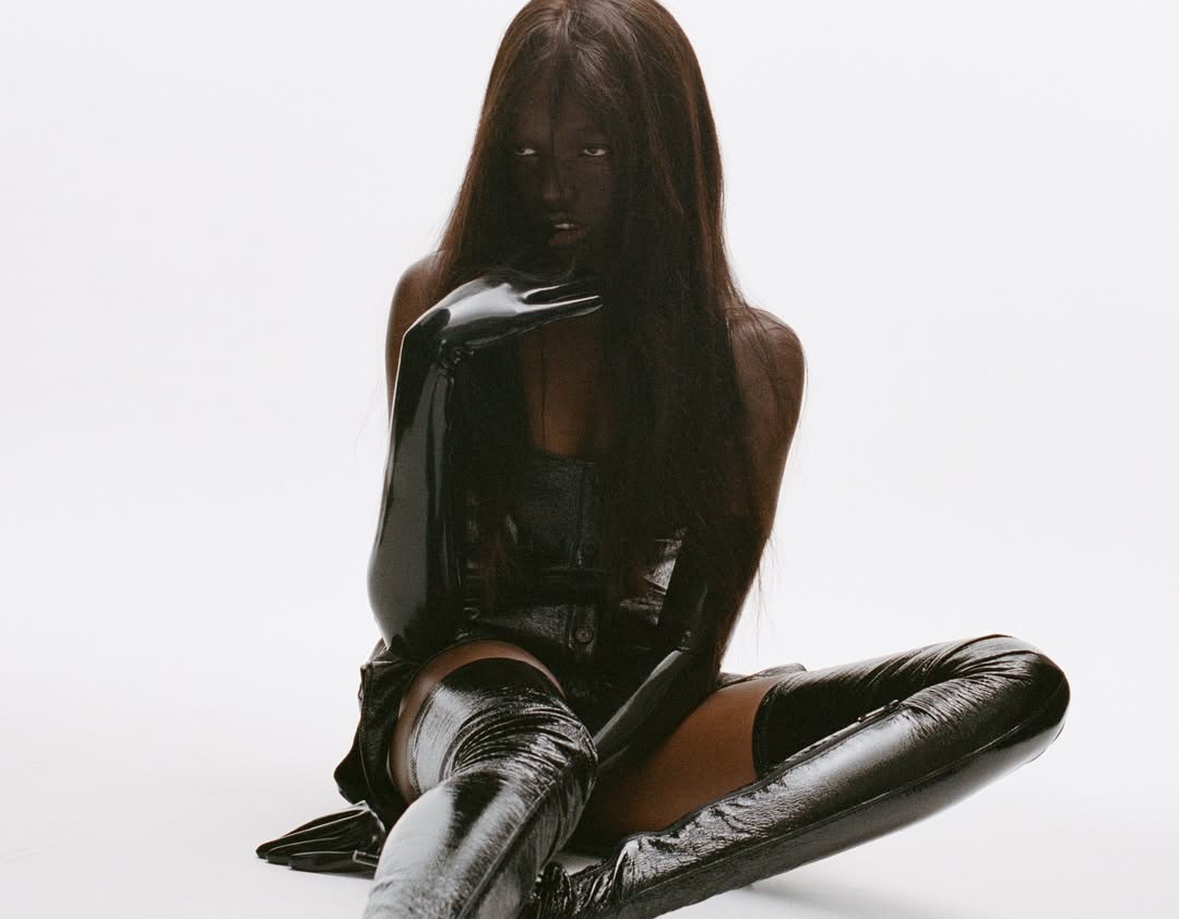 Woman in a black latex bodysuit and thigh-high boots sits on a white surface, with matching black latex gloves and dark hair.