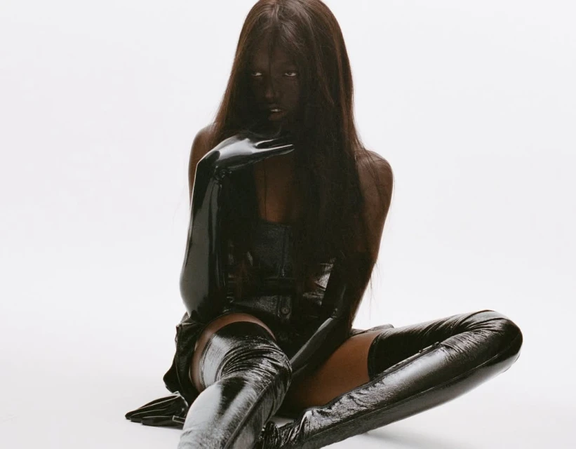 Woman in a black latex bodysuit and thigh-high boots sits on a white surface, with matching black latex gloves and dark hair.