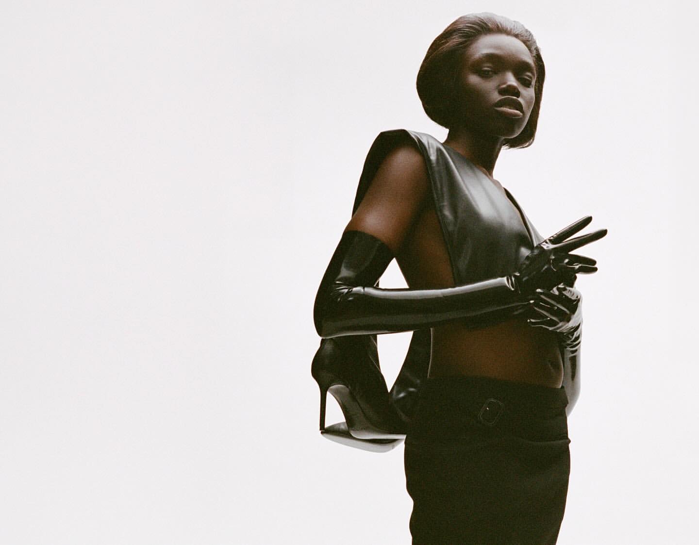 Striking fashion shot of a Black female model posing in dark sleeveless top and gloves, high heels, and pants against a white backdrop.