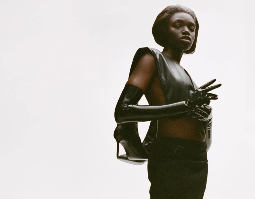 Striking fashion shot of a Black female model posing in dark sleeveless top and gloves, high heels, and pants against a white backdrop.