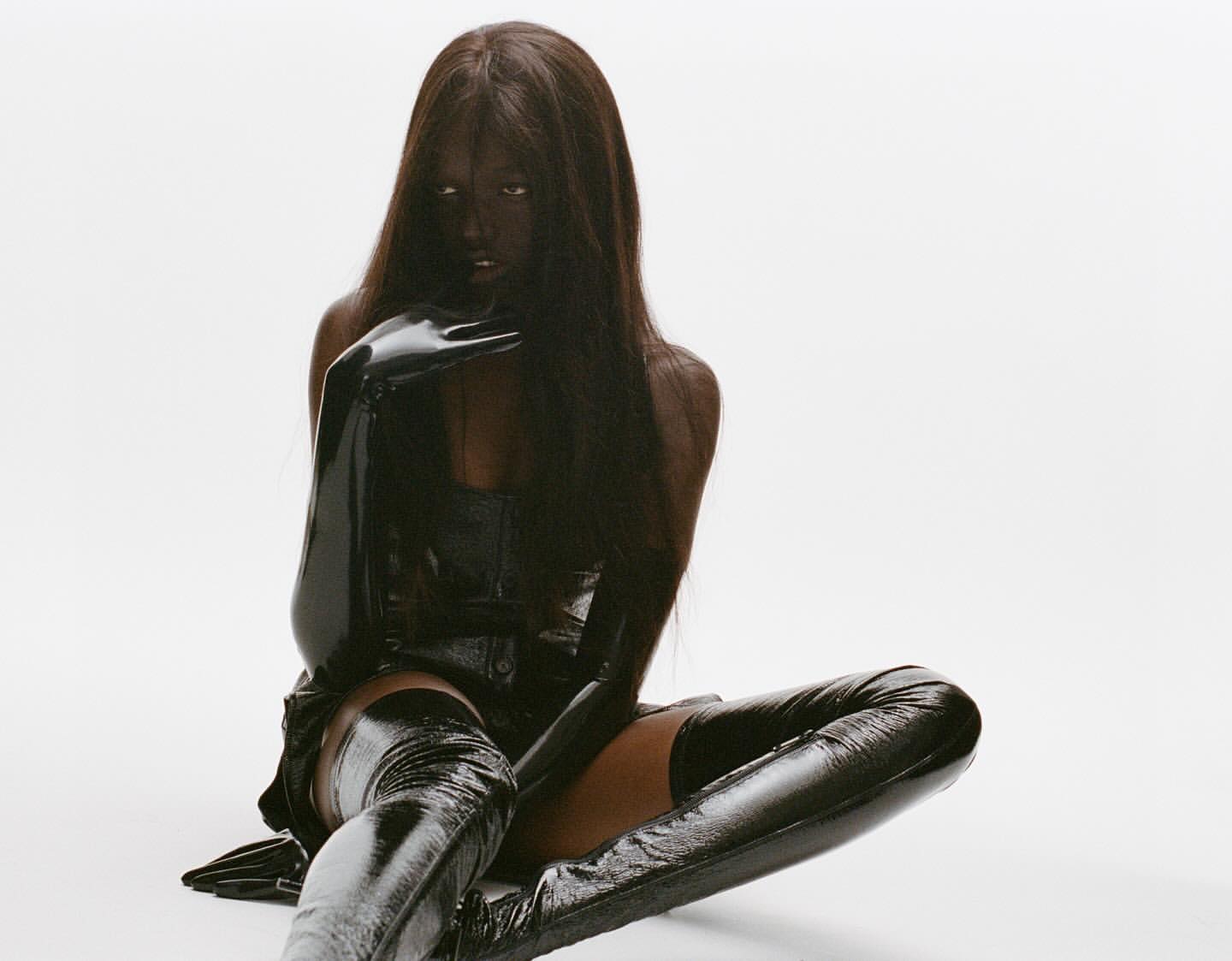 Woman in a glossy black outfit, including a corset, gloves, and thigh-high boots, poses in a studio against a stark white backdrop.